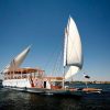 A traditional sailboat with white sails glides on a calm river under a clear blue sky.