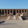 Ancient Egyptian temple ruins with a central staircase leading to a columned entrance under a clear sky.