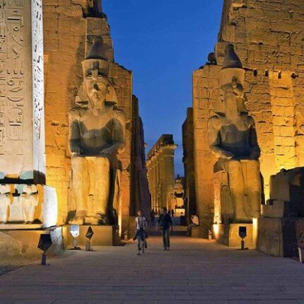 Two colossal statues of pharaohs illuminated at night at the entrance to an ancient Egyptian temple.