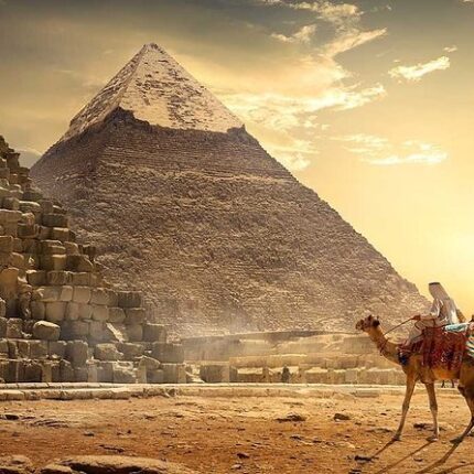 A person riding a camel in front of a pyramid at sunset, casting a golden glow on the scene.