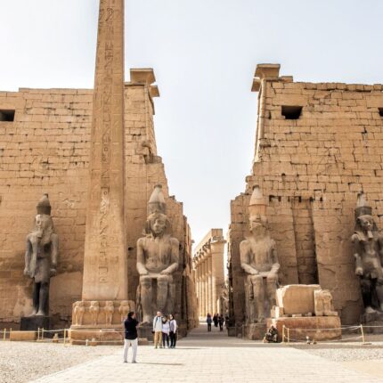 Tourists exploring the entrance of Luxor Temple, flanked by statues and an obelisk under a clear sky.