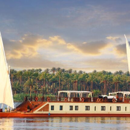 A traditional Nile riverboat with large sails cruising past a lush, palm-lined riverbank.