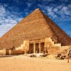 The Great Pyramid of Giza with its entrance and surrounding ruins under a blue sky with scattered clouds.