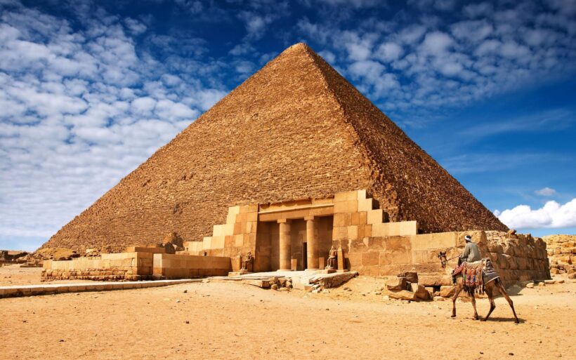 The Great Pyramid of Giza with its entrance and surrounding ruins under a blue sky with scattered clouds.