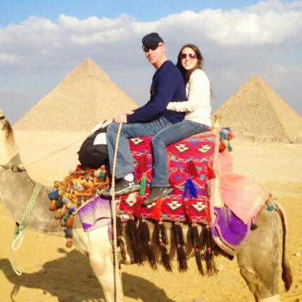 A couple riding a decorated camel with the Pyramids of Giza in the background under a partly cloudy sky.