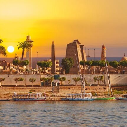 Sunset view of Luxor with boats on the Nile River and ancient structures silhouetted against the sky.