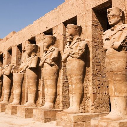 Row of ancient statues of Ramses II at the Karnak Temple complex under a clear sky.